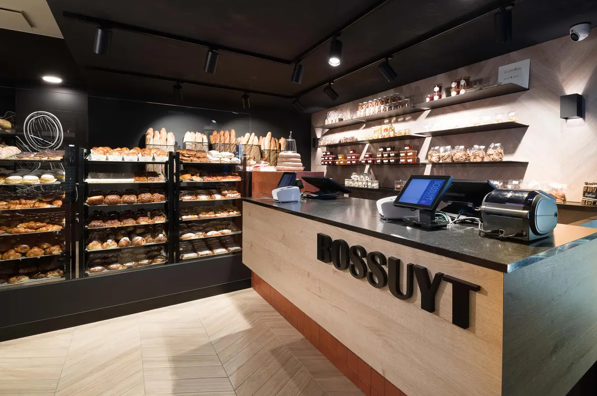 De toonbank van Bakkerij Bossuyt met rekken vol brood, ontbijtkoeken en koffiekoeken, en schappen met confituur en koekjes